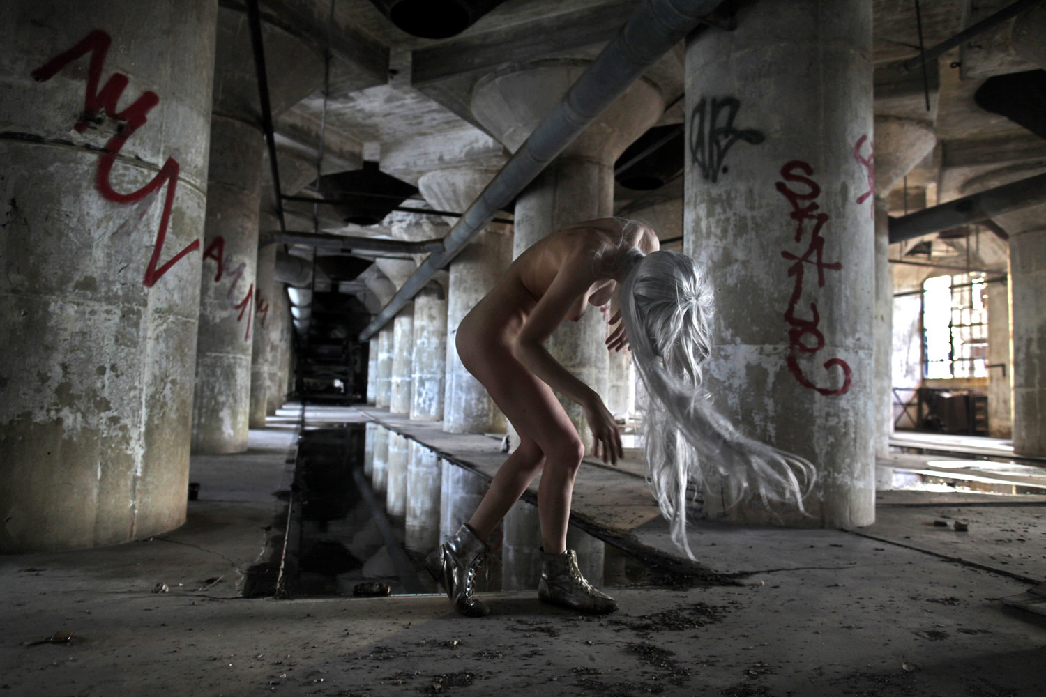 A pale-haired performer bends forward in a dim concrete interior with columns, puddles, and graffiti.